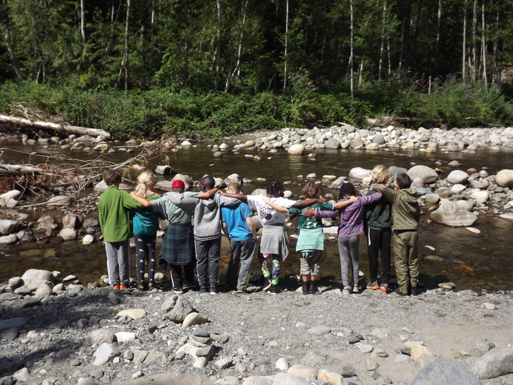 Campers stand arm-in-arm together in front of the river, their back to the camera