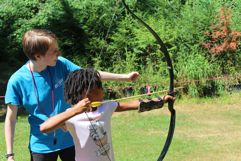 An action shot of a staff member helping a young camper shoot a bow and arrow